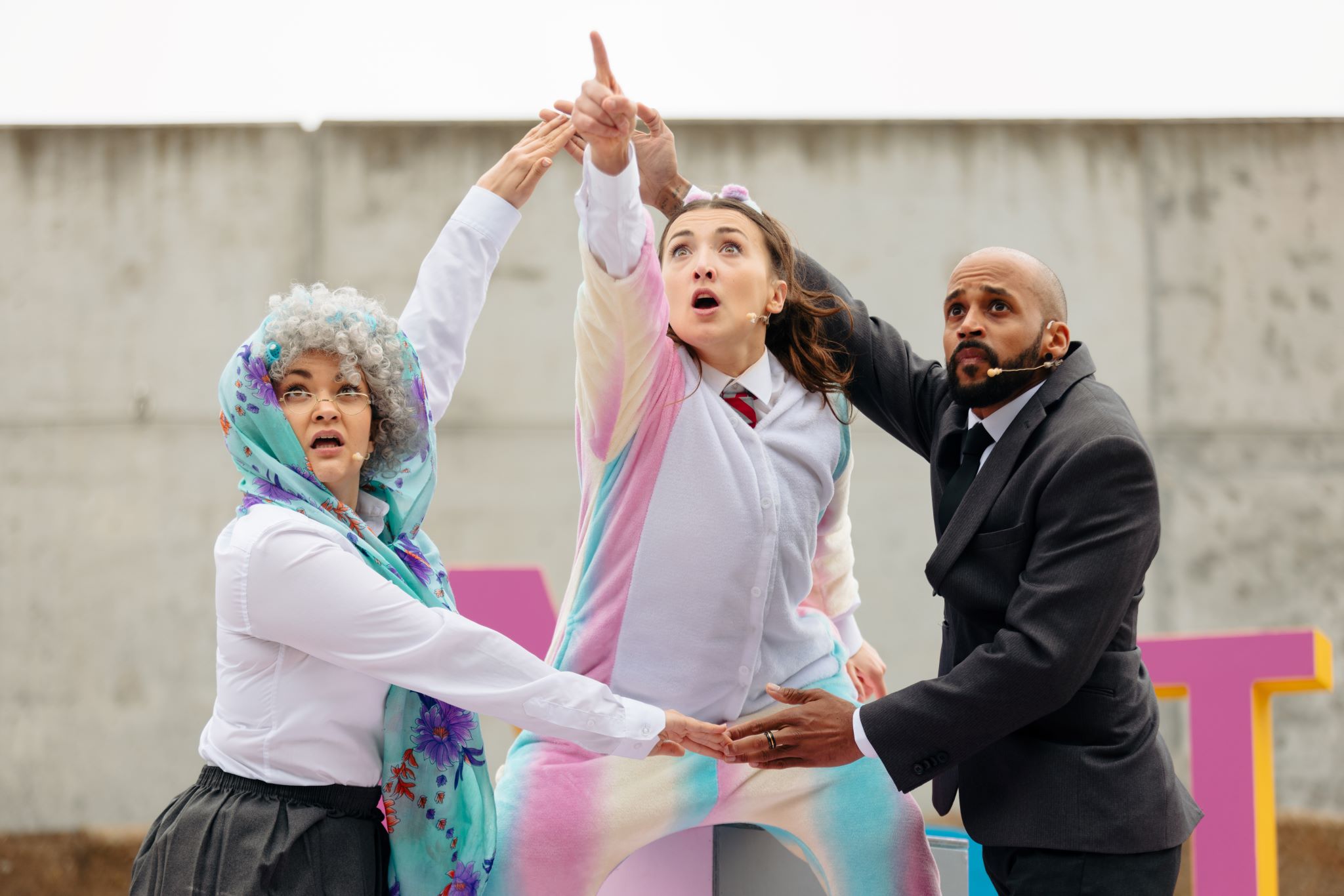 A performer wearing a multicoloured pastel rainbow unicorn onesie over a school uniform points up in the distance. Two other performers form a circle with their arms around her. The one to her left is wearing a black suit; the one to her right is wearing a curly grey wig and blue scarf over her head.