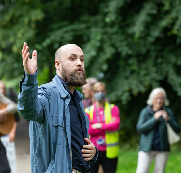 Leigh Johnstone, Artistic Director of All in the Mind Festival is stood at the festival speaking, his arm raised in the air