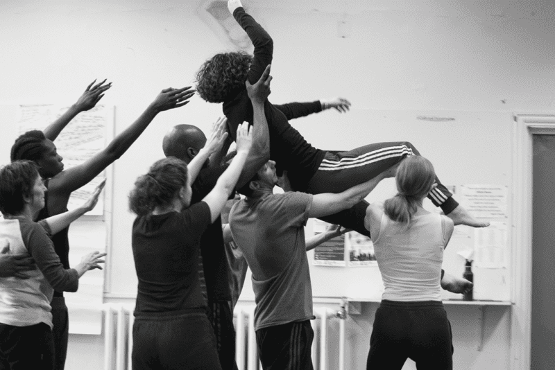 A black-and-white photo from rehearsals. A performer is being lifted up by four other performers, with three performers spotting behind them.