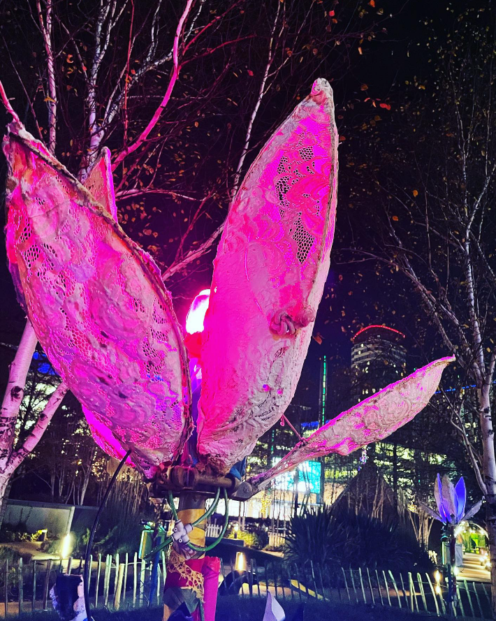 Outdoors at night, a big robotic flower with petals made out of lace and bent metal wire is lit by a pink light.